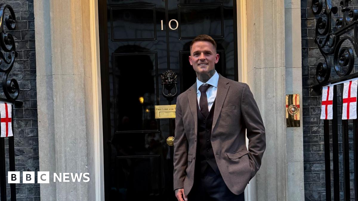 A smiling man stood outside the front door of Number 10 Downing Street. He is wearing a brown suit jacket with black waistcoat underneath, white shirt and brown tie. Attached to some black railings are St George's flags.