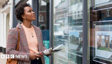 A stock image of a lady perusing an estate agent's available properties in the window. She carries a newspaper, a handbag, and a blazer.