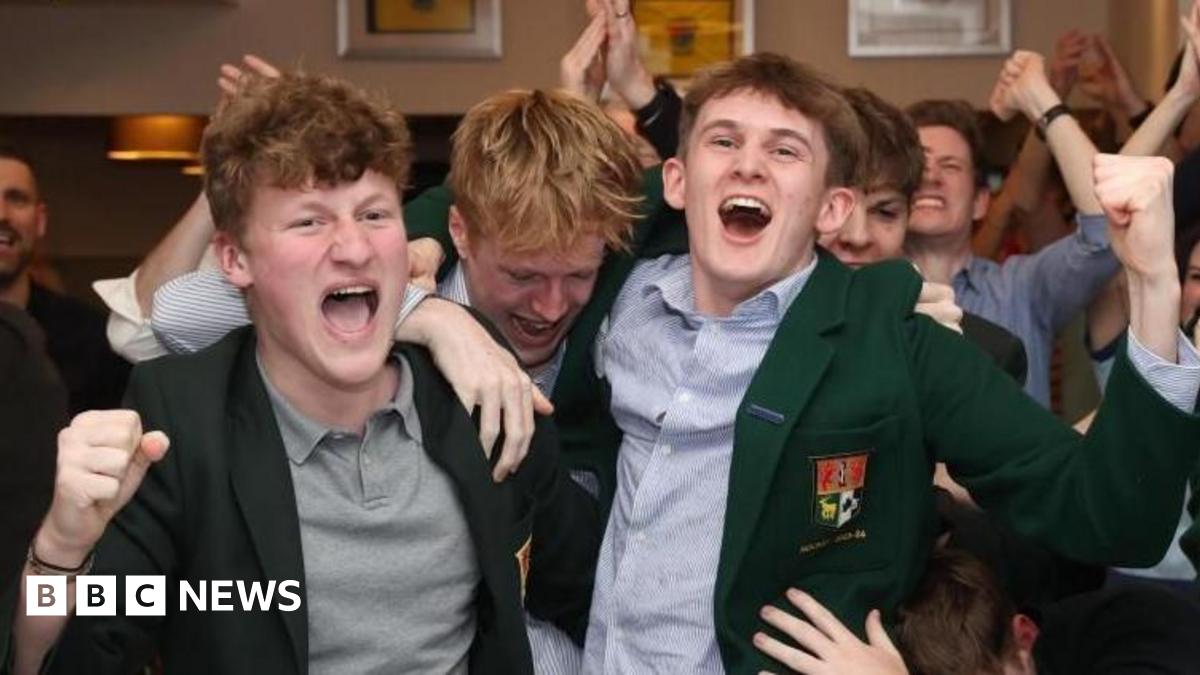 A group of young people sit at the front of a large crowd in a golf club supporting Rory McIlroy. They are all wearing blue shirts and the two young men in the middle are also wearing dark green golf club blazers. Drinks sit on the table. The people are all cheering and punching the air.