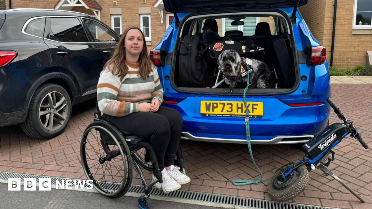 Kay Millar is sitting in a wheel chair next to a blue car with the boot open. Her dog is standing in the boot.
