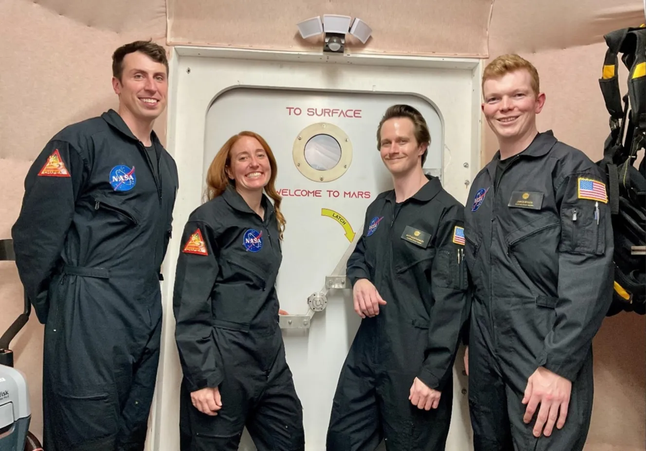 The four CHAPEA Mission 2 crew members, Ross Elder, Ellen Ellis, Matthew Montgomery, and James Spicer, pose in front of a door marked "TO SURFACE" and "WELCOME TO MARS" inside the CHAPEA habitat.