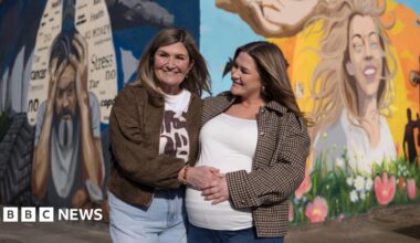 Wendy Robinson pictured with her daughter Amy standing in front of a colourful mural on two sides of a wall. Amy, on the right, has blonde hair and is smiling. There are blue skies, sun, flowers and birds behind her. On the mural towards the left is a picture of a man holding his head in his hands with words including 'cancer, stress, no, health' written in a lung-diagram on a dark blue background.
