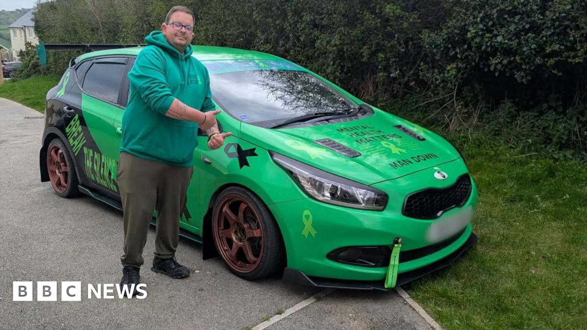 A man with brown hair and glasses and wearing a green hoodie, dark green trousers with black boots is stood in front of his bright green car. He has his thumbs up and is looking straight at the camera.