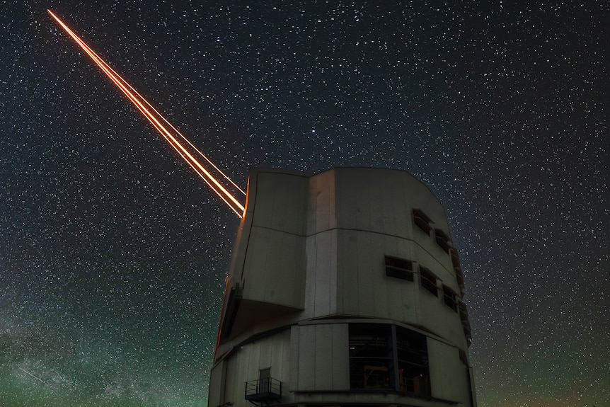 Grey cylindrical building shoots three orange lasers into a starry night sky.