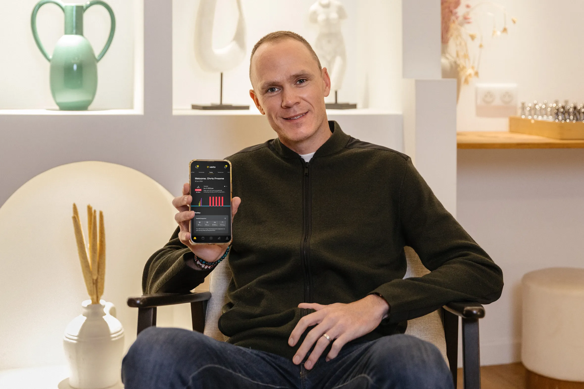 Chris Froome sitting in a chair, holding a smartphone displaying the Vekta app.