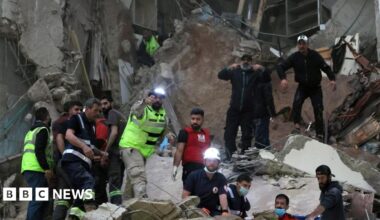 A number of rescue workers, some with helmets and head torches, others with face masks, work in the rubble of a destroyed building