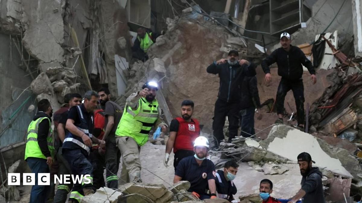 A number of rescue workers, some with helmets and head torches, others with face masks, work in the rubble of a destroyed building