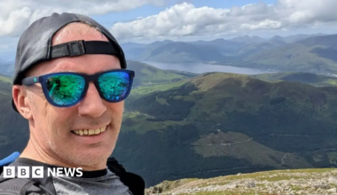 A man wearing green-tined sunglasses, a black cap on backwards and a grey T-shirt is standing on a mountain. In the background is a view of rolling hills and a reservoir.