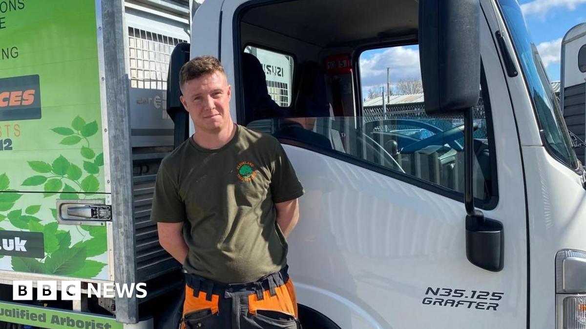 Jacob Francis standing in front of the driver's door of a large white van. His hands are behind his back. His hair is short and brown and he is wearing a khaki t-shirt.