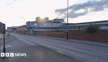 A wide, empty street in an urban area under a partly cloudy sky. The road curves gently and has clear lane markings, with pavements and signposts along the edges. Brick walls and industrial-style buildings line the right side of the street, while trees and smaller buildings appear on the left.