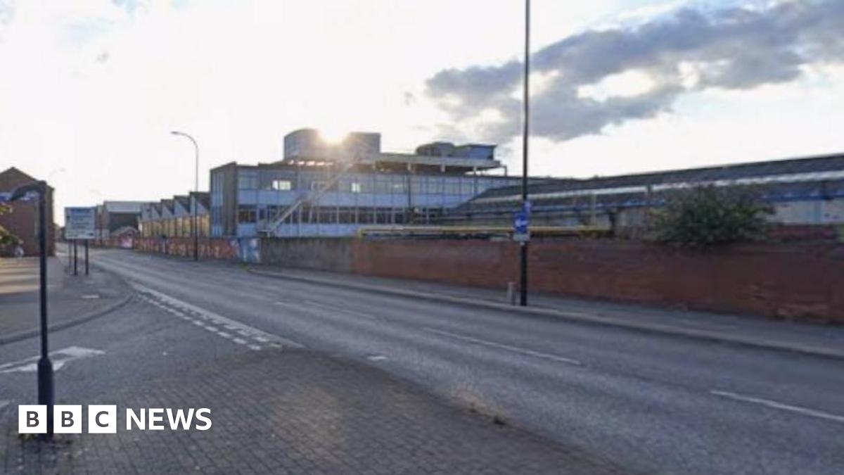 A wide, empty street in an urban area under a partly cloudy sky. The road curves gently and has clear lane markings, with pavements and signposts along the edges. Brick walls and industrial-style buildings line the right side of the street, while trees and smaller buildings appear on the left.