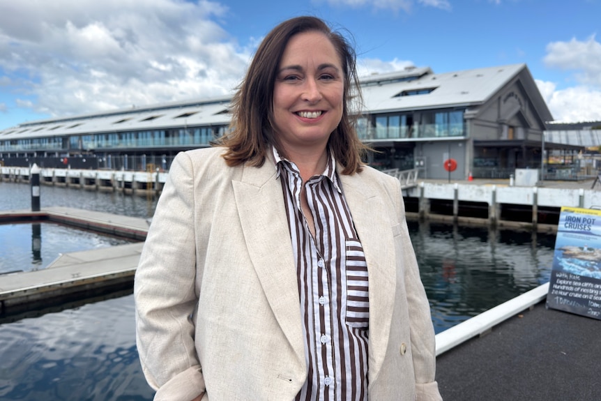 A woman with brown hair and white blazer stands in front of a long building on a pier.