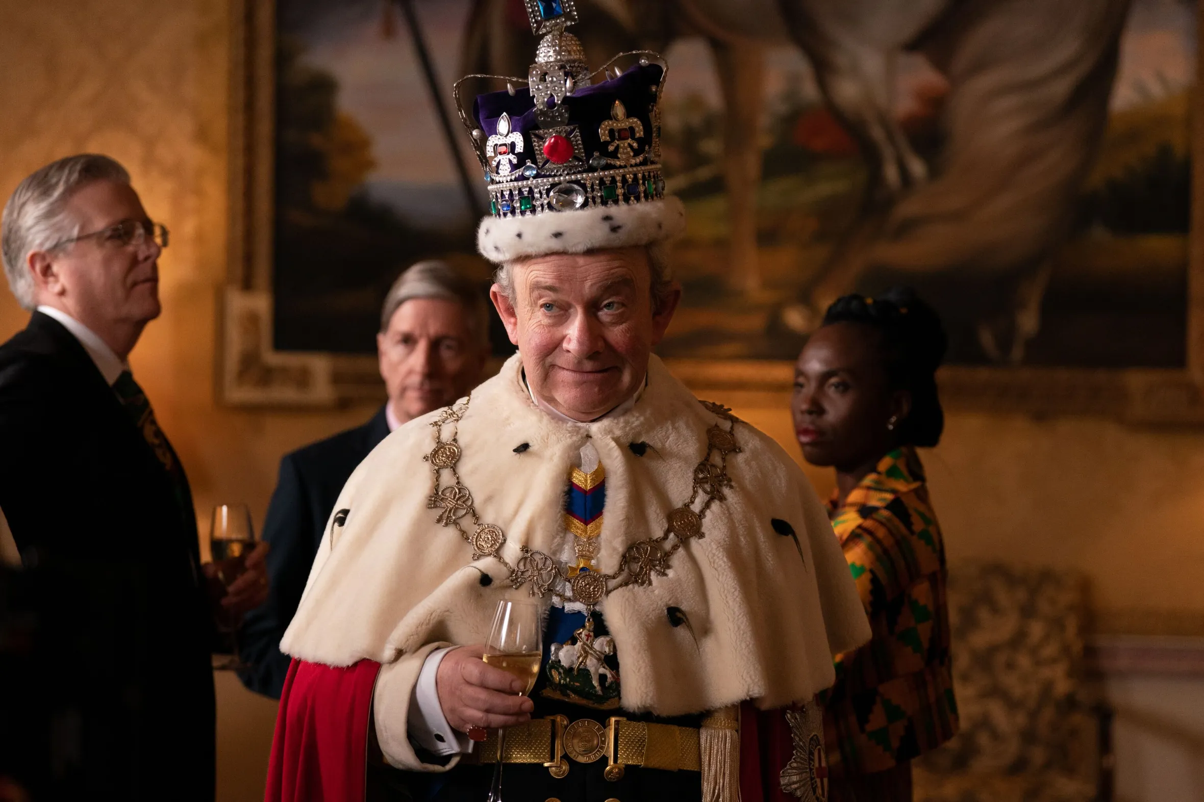 Harry Enfield as Charles in a crown, ermine, and regalia, holding a glass of wine.