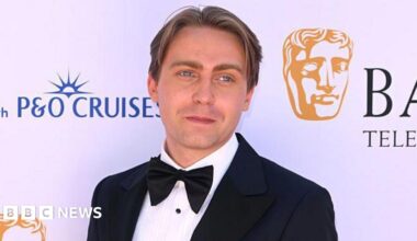 Barney poses on a red carpet, in front of a white backdrop with with Bafta branding on it. He has dark blonde hair, and wears a black tuxedo with a black bow tie and a white shirt.