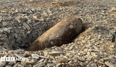 The picture shows a large, rusted cylindrical object, a a German SC250 air-dropped bomb, lying on its side in a shallow pit at what appears to be a construction or excavation site. The object is partially embedded in loose grey rubble made up of broken stone and aggregate.