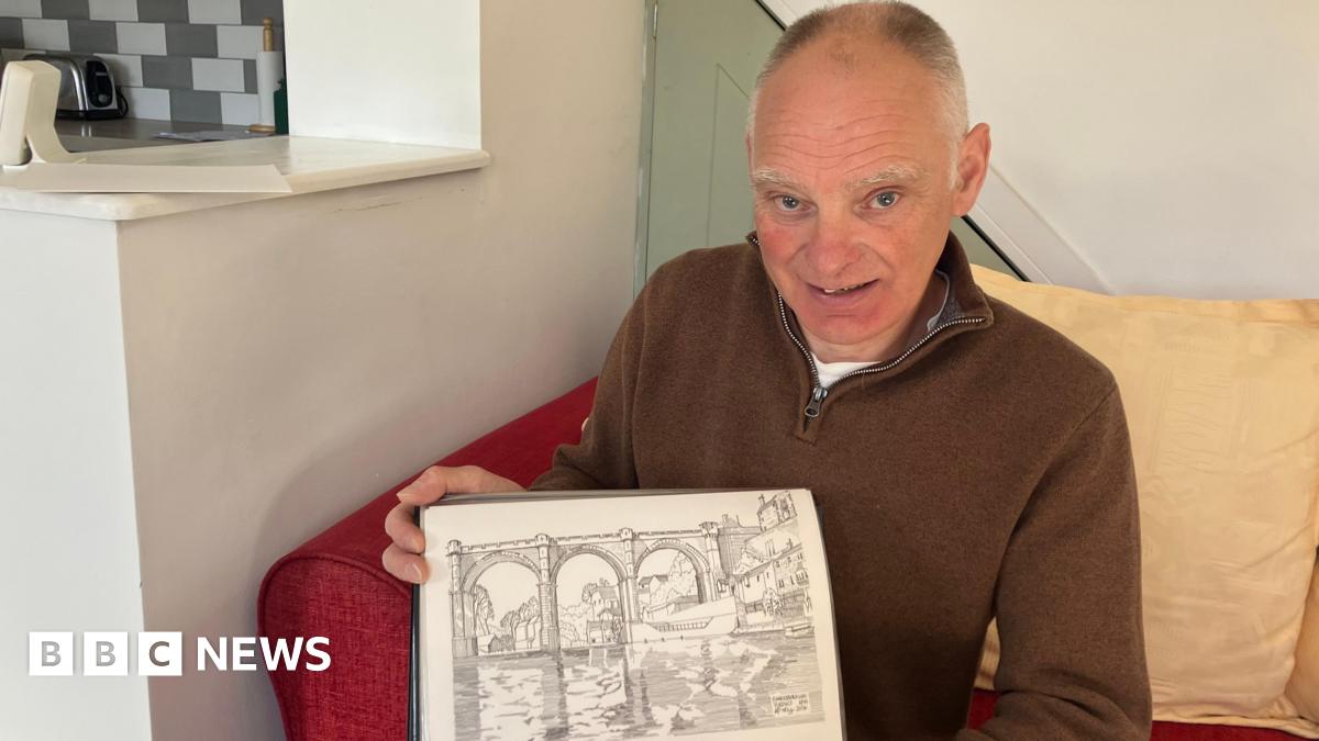 A man is sitting on a red sofa with a yellow cushion behind him holding a hand drawn picture of Knaresborough Viaduct