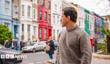 Young man in a jumper looks back over his shoulder at a row of brightly painted terraced Notting Hill townhouses.