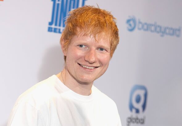 An individual with red hair is standing in front of a wall adorned with logos, wearing a white t-shirt and smiling at the camera An individual with red hair is standing in front of a wall adorned with logos, wearing a white t-shirt and smiling at the camera