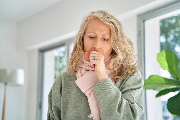 An adult female with blonde hair, wearing a green long-sleeved top, appears to be coughing while standing in a room with glass w