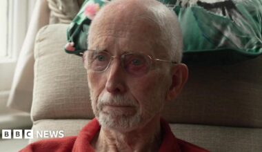 Brian Gundry, is a resident of a residential home in Dorchester. He is sittings in a chair in his room. There is a cushion behind his head. He's wearing a red shirt and has glasses and a beard.