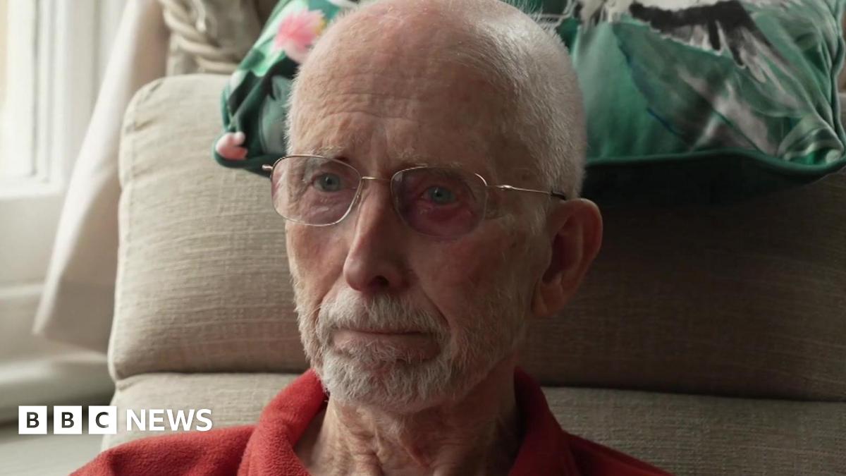 Brian Gundry, is a resident of a residential home in Dorchester. He is sittings in a chair in his room. There is a cushion behind his head. He's wearing a red shirt and has glasses and a beard.