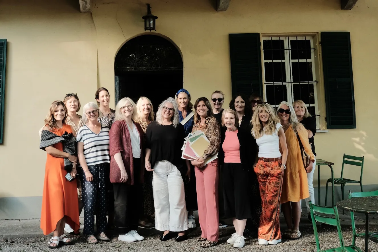 A group of 18 women posing together in front of a light yellow building.
