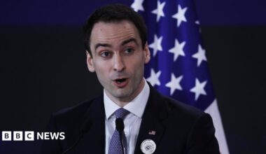 Michael Kratsios, a White House director and advisor on technology, speaking into a microphone at a podium, wearing a black suit jacket, white dress shirt and blue patterned neck tie. An American flag is positioned upright behind him.