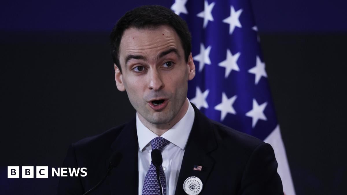 Michael Kratsios, a White House director and advisor on technology, speaking into a microphone at a podium, wearing a black suit jacket, white dress shirt and blue patterned neck tie. An American flag is positioned upright behind him.