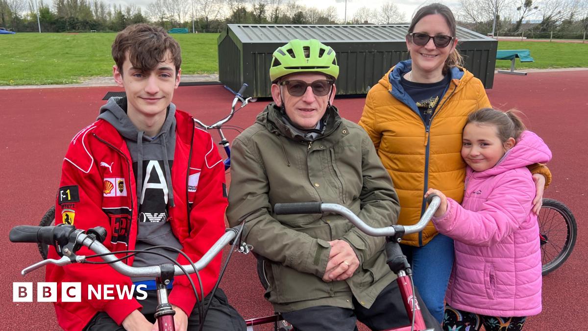 An old man sits on an adapted bike next to his 20-year-old grandson. His daughter and granddaughter are stood beside him, hugging. The man is wearing a helmet and the family are all wearing coats. They are at an athletics track.