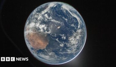 A picture of the Earth from space, which centres the planet against the dark background of space. It's a round blue planet. Clouds can be seen and a thin green aurora at the top.