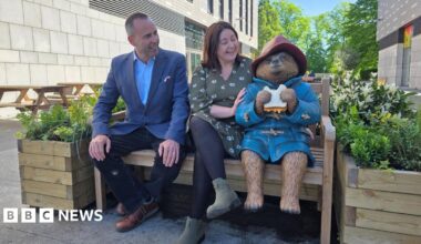 Paddington Bear statue dressed in his trademark red hat and blue coat while eating a marmalade sandwich. He is sitting on a bench with two wooden planters filled with flowers beside him. A man dressed in a blue suit and a woman in a green dress sit beside Paddington