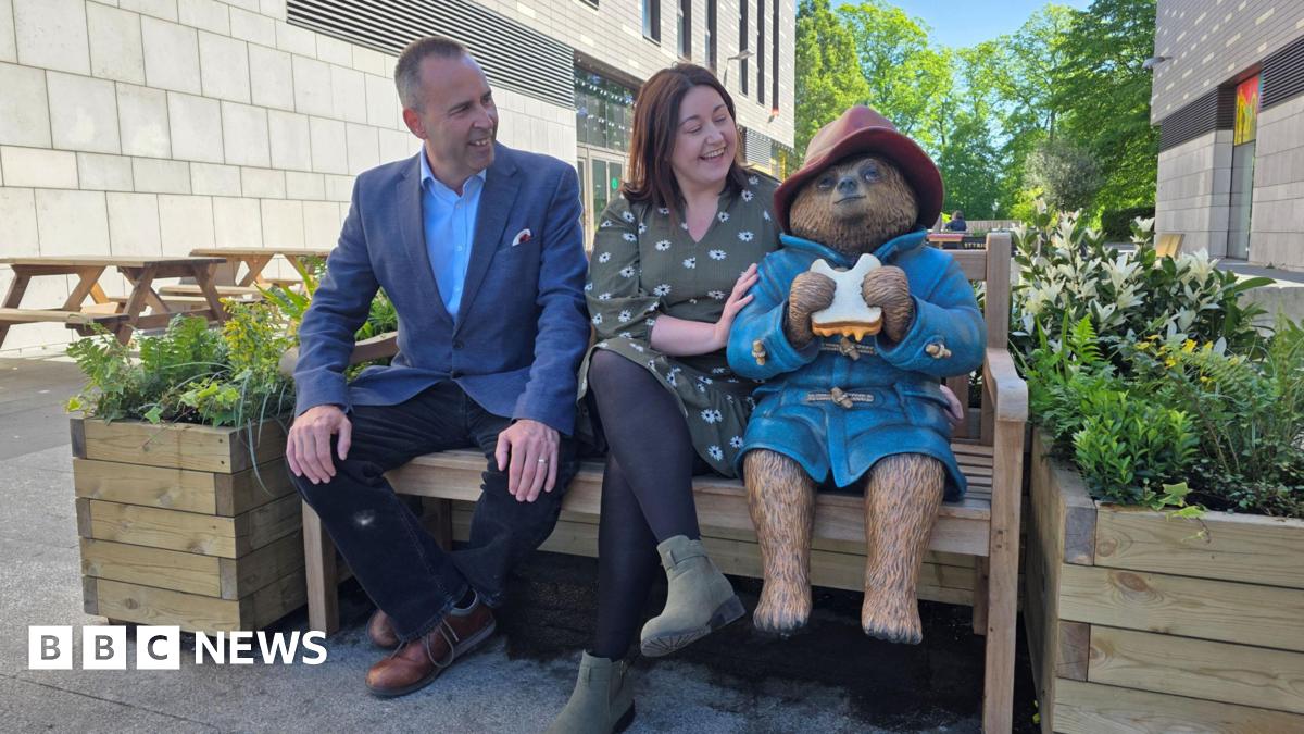 Paddington Bear statue dressed in his trademark red hat and blue coat while eating a marmalade sandwich. He is sitting on a bench with two wooden planters filled with flowers beside him. A man dressed in a blue suit and a woman in a green dress sit beside Paddington