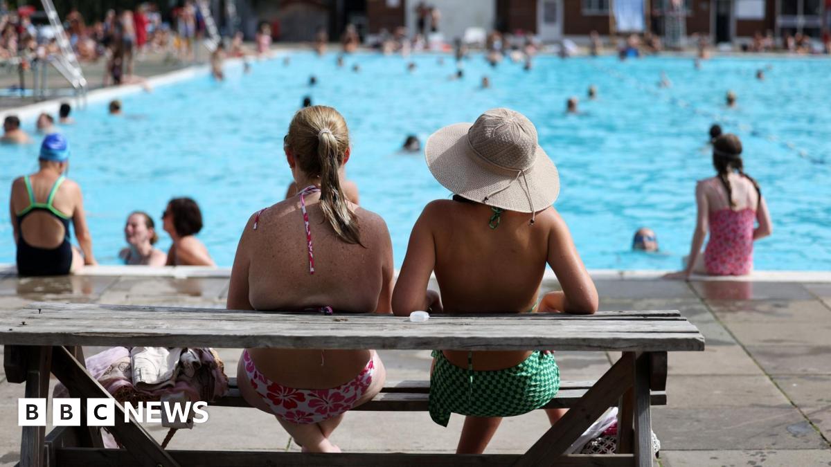 The back of two women sat at a wooden bench, looking out over a busy outdoor swimming pool. One of them is wearing a hat and it is very sunny.