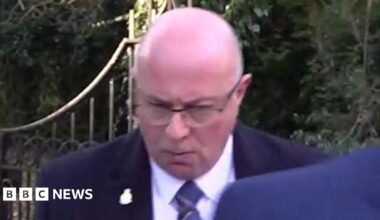 PC Mark Roberts leaving Durham Crown Court. He is wearing a dark suit and a blue striped tie. He is bald, wearing glasses and looking down.