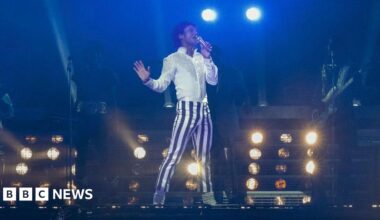 A film still showing a young man striking a pose on stage as he sings into a microphone. He has black hair in a mullet cut and wears a bright white shirt and white and blue striped trousers, which make him stand out against a dark background. The lighting is low and the shapes of his backing musicians can only just be made out. Stage lights shine in the background.