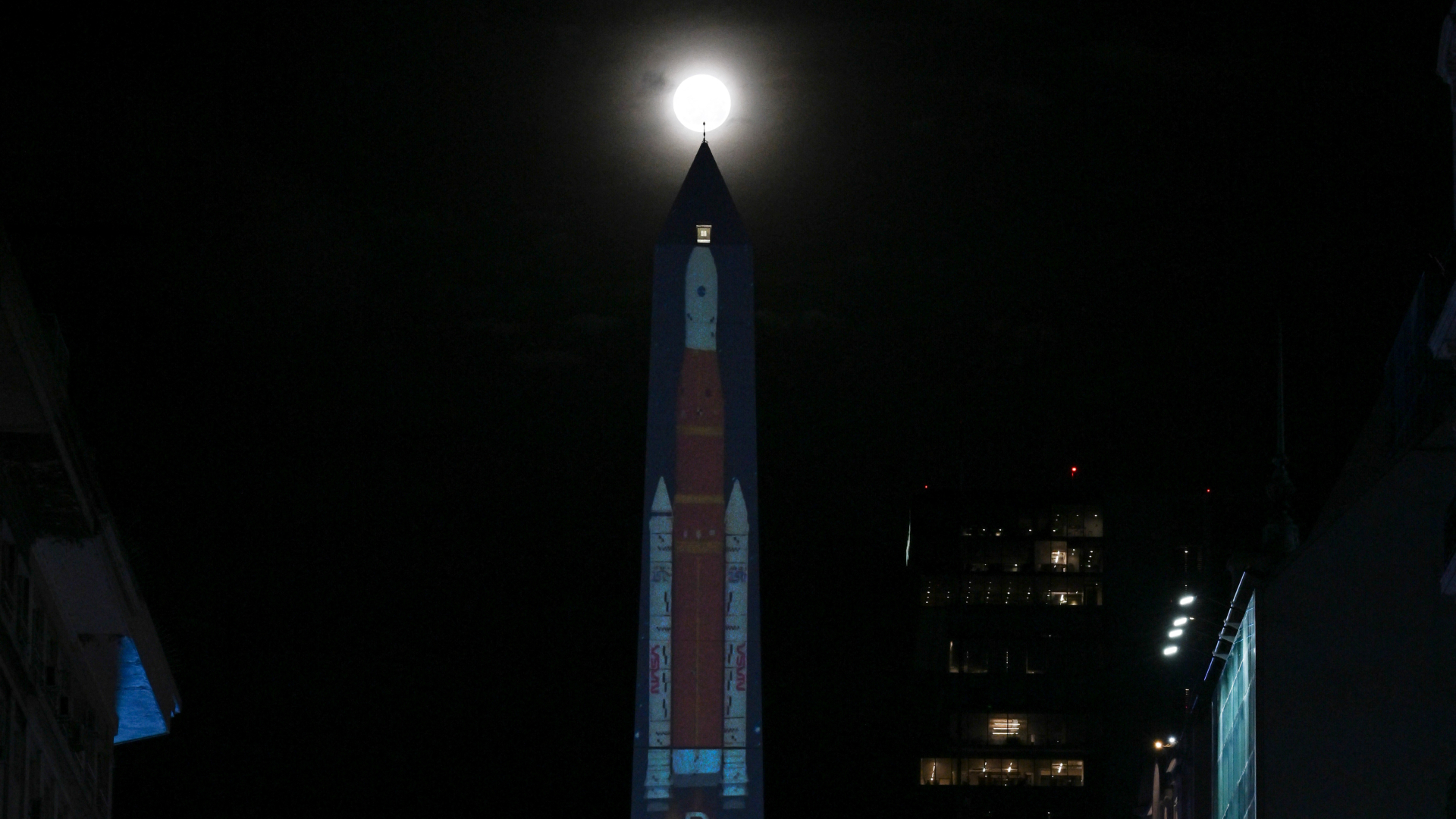 A large orange and white rocket is projected on the side of an obelisk at night surrounded by buildings. The moon is visible glowing above the monument.