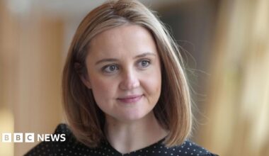A woman with shoulder-length blonde hair, looking off camera. She is wearing a black and white polka dot top.