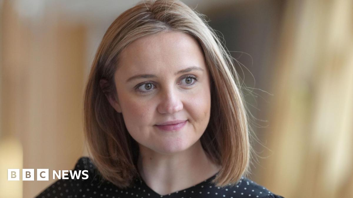 A woman with shoulder-length blonde hair, looking off camera. She is wearing a black and white polka dot top.