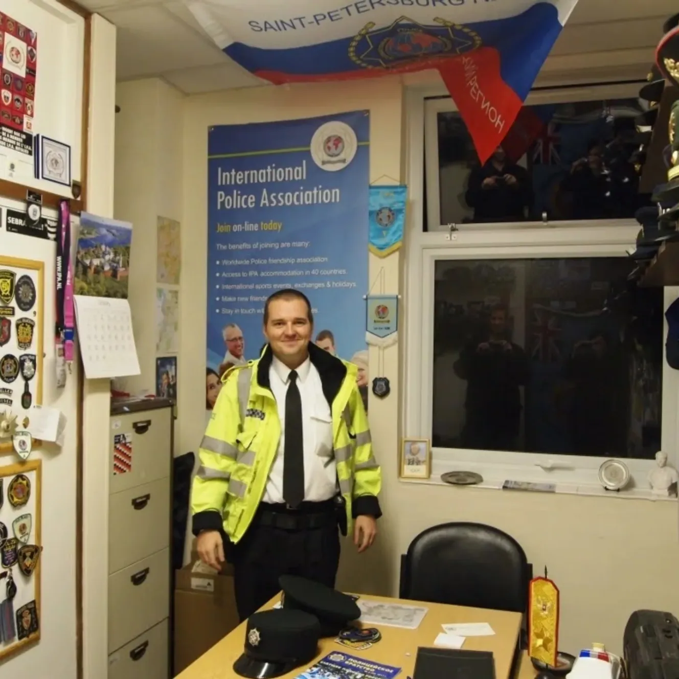 Mark Bullen, a former British police officer, stands in an office with a Russian flag above him and an "International Police Association" poster behind him.