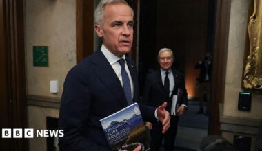 An image of Mark Carney in the foyer of Canada's House of Commons. He is seen speaking to a person off-camera while holding a document titled 'Un Canada Fort Pour Tous'. Beside him is finance minister Francois-Philippe Champagne, who is looking over at Carney and is holding the same document.