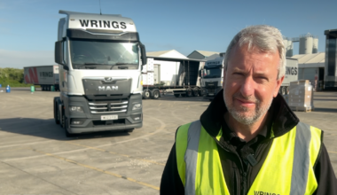 A man stood in a yard with a large white lorry behind him. He is wearing a reflective yellow jacket. He has short grey hair and a short beard.