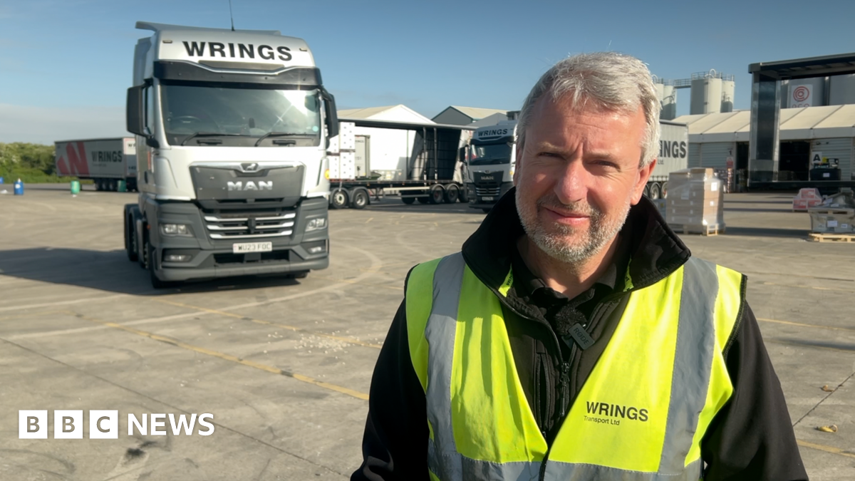 A man stood in a yard with a large white lorry behind him. He is wearing a reflective yellow jacket. He has short grey hair and a short beard.