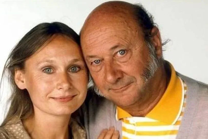 A portrait of a mature couple standing closely together, both individuals are smiling and posing for the camera against a plain background. The woman has long, dark hair, and the man has a yellow shirt with white stripes and a beard.