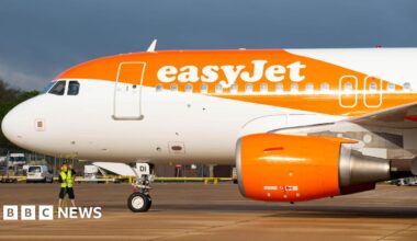 The front of a plane parked at an airport, branded with the word easyJet, with a streak of orange paint over the body of the plane. A person in a high-vis vest is standing under the plane.
