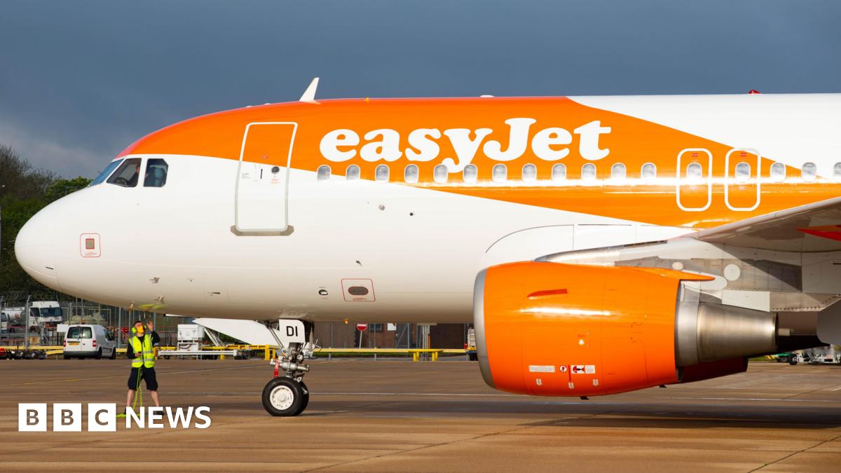 The front of a plane parked at an airport, branded with the word easyJet, with a streak of orange paint over the body of the plane. A person in a high-vis vest is standing under the plane.