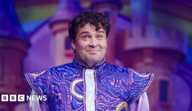 Joe McElderry is on stage and smiling sheepishly. He has short, curly, dark hair. He is wearing a costume which is gold and purple and has pointy shoulders.