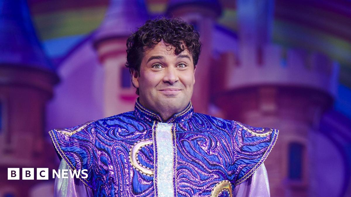 Joe McElderry is on stage and smiling sheepishly. He has short, curly, dark hair. He is wearing a costume which is gold and purple and has pointy shoulders.