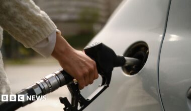 A woman using a gas pump to refuel vehicle during energy crisis. She has a silver car, and is wearing a white sweater.
