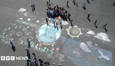 An ariel view of the artwork on Plymouth Hoe. There is Earth at the centre, with 'Happy 100 Birthday Sir David Attenborough' on it. There is a whale to Earth's right and trees and mountains to its left. A choir is gathered at the top the artwork, with people lined up taking photos on the other side.