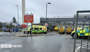 Ambulances parked in a car park outside of a hospital. The hospital building is large and grey. A large red sign is displayed in front of the car park which reads 'emergency vehicles only beyond this point'.